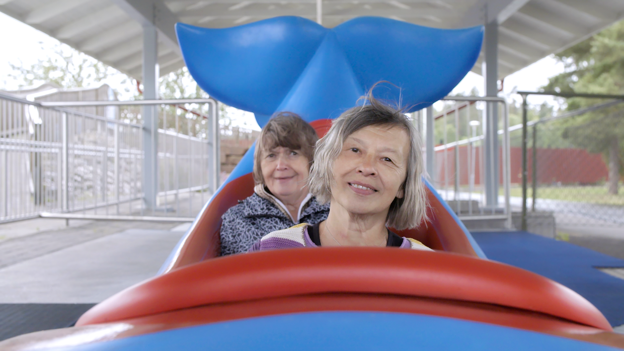 Two older women sit in an amusement park ride shaped like a whale, only the tail is visible behind them as they smile.