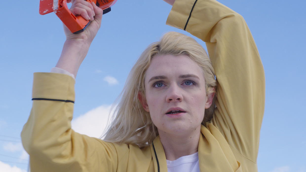 A young women with platinum blond hair holds something out of sight above her head as she gazes in front of her. She is wearing a yellow long-sleeve button-up top.