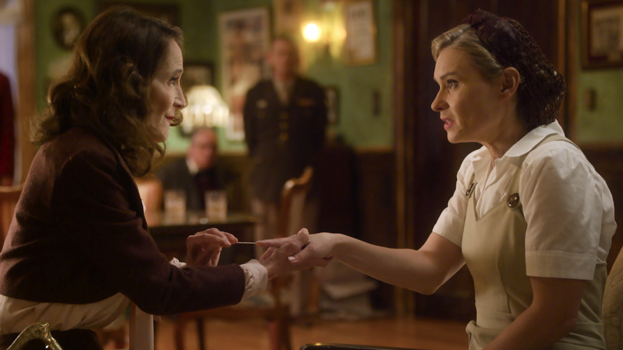 Two women sit in a '40s-era style parlor room, one woman wearing a military uniform is holding a long needle against the finger of the other woman, who is blonde and wears beige overalls and a white linen shirt.