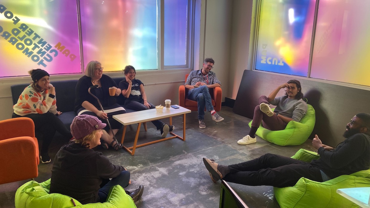 A diverse group of people sit in neon green beanbag chairs in a circle in a room, the window reads PAM CUT: Center for an Untold Tomorrow.