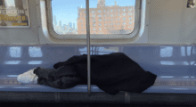 A man sleeps with a jacket covering his face and body on a vacant row of subway seats, Lower East Side housing projects appear outside of the window behind him.
