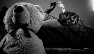 A black and white photo of a woman laying on her back and blowing out a plume of smoke while a big teddy bear sits to the left and in the forefront of the frame.