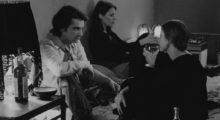 Three people, one man and two women, sit on a bed drinking wine and looking off in different directions in this black and white film still.