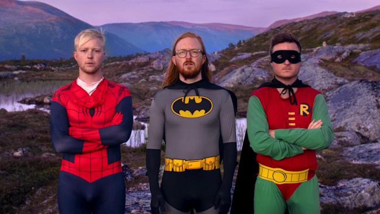 Three grown men stand in front of an image of mountains, they don, from left to right, three superhero costumes: spiderman, batman and robin.