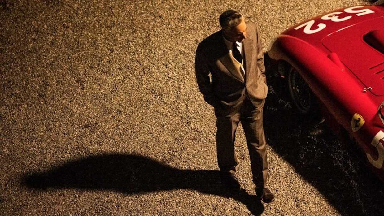 A man wearing a brown suit walks beside a bright red Ferrari.