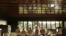 A group of chefs stand in an assembly line in a huge French kitchen, the wall that they face features floor to ceiling windows that display a verdant view.