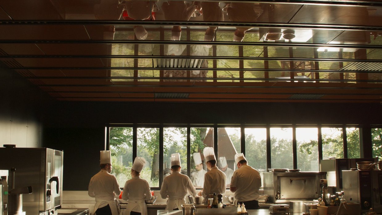 A group of chefs stand in an assembly line in a huge French kitchen, the wall that they face features floor to ceiling windows that display a verdant view.