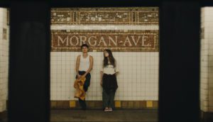 A trans man with cropped brown hair and a teenage girl with straight, long brown hair stand at the Morgan Ave L stop. They both wear white shirts and black pants.