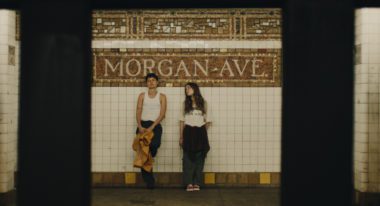 A trans man with cropped brown hair and a teenage girl with straight, long brown hair stand at the Morgan Ave L stop. They both wear white shirts and black pants.
