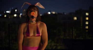 A woman wears a pink bikini top, a ball gag, bunny ears and a pig's nose outside at night.