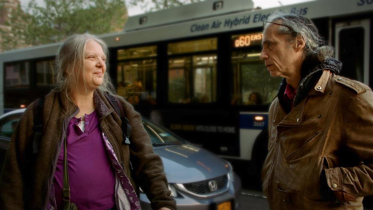 An older man and woman stand on a sidewalk as a Q60 bus passes by them in Queens.