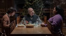 A teenage boy, elderly white man and middle-aged Black woman sit at a wooden dining table with a Christmas dinner.