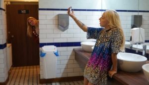 A blonde woman wearing a sparkly dress made up of silver, blue and purple sequins takes a selfie in a bathroom. Another blonde woman peeks around a corner and watches her.