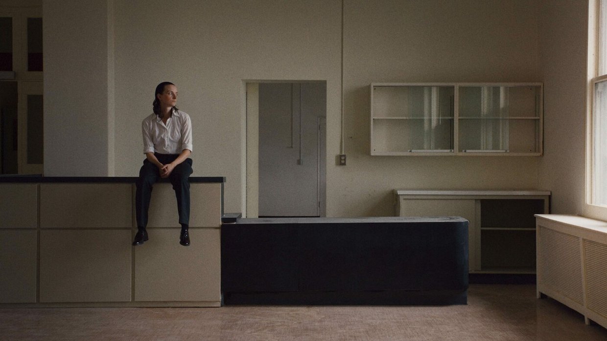 A woman wearing a white shirt and black pants sits on a counter in a completely empty room. She looks to her left at something out of frame.