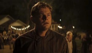 A man in '20s-era garb stands amid a sparse crowd at night, his jaw slack and eyes wide in reaction to something happening off-screen.