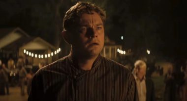 A man in '20s-era garb stands amid a sparse crowd at night, his jaw slack and eyes wide in reaction to something happening off-screen.