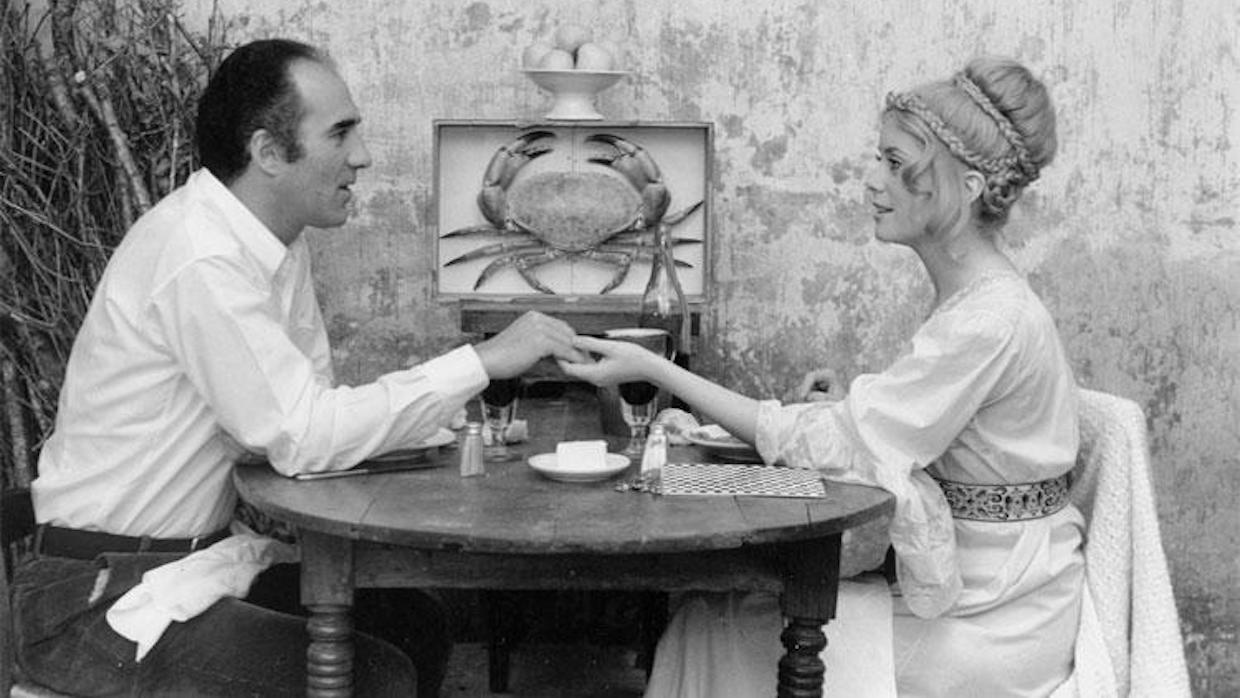 A black and white image of a man and a woman sitting at an outdoor table with a framed crab hung between them. The man and woman hold hands at the center of the table.