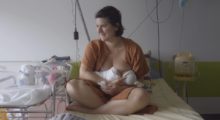 A woman with short brown hair and an orange hospital gown breastfeeds her newborn baby in the hospital.