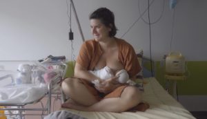 A woman with short brown hair and an orange hospital gown breastfeeds her newborn baby in the hospital.