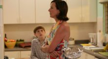 A mother in a color apron and her young son in a suburban kitchen.