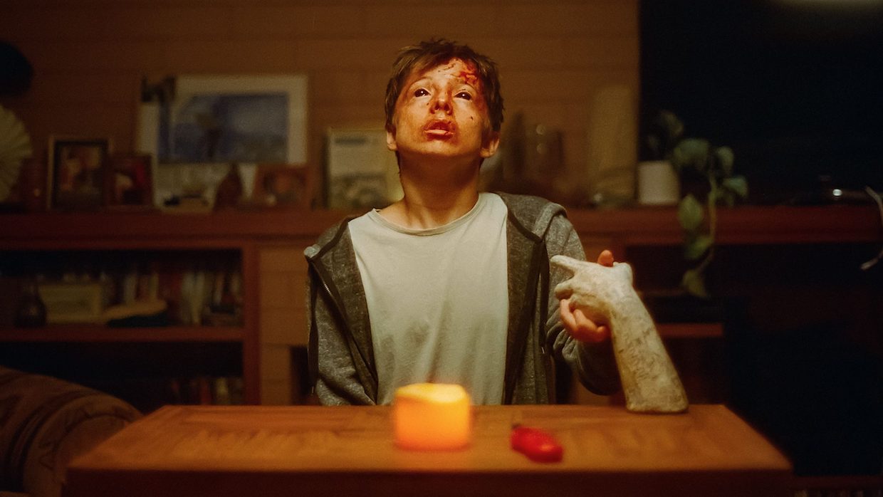 A pre-teen boy sits at a wooden table with one candle lit. He holds a ceramic hand. His face is bloody and his eyes are black.