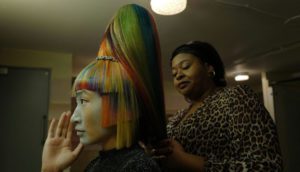 A woman with long, rainbow-colored hair in a tiered high ponytail sits at a salon chair with another woman wearing cheetah print and tightly coiffed hair works on the other woman's hair.