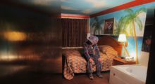 A man wearing a suit with a retro '90s carpet design tilts his head down as he sits in a tropical-themed motel room. His 10-gallon cowboy hat covers his face as he gazes downward.