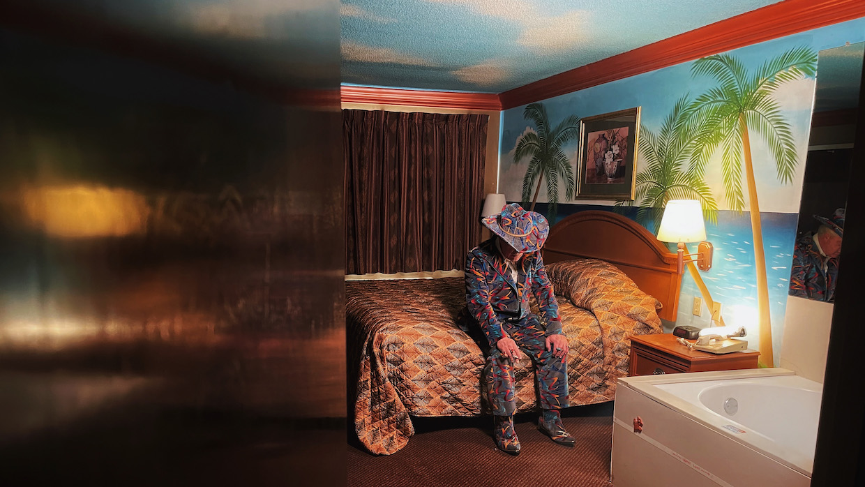 A man wearing a suit with a retro '90s carpet design tilts his head down as he sits in a tropical-themed motel room. His 10-gallon cowboy hat covers his face as he gazes downward.