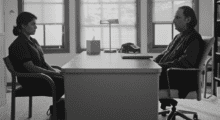 A black and white shot of a young woman sitting across from a middle-aged man in his office. He is her therapist and she is his patient.