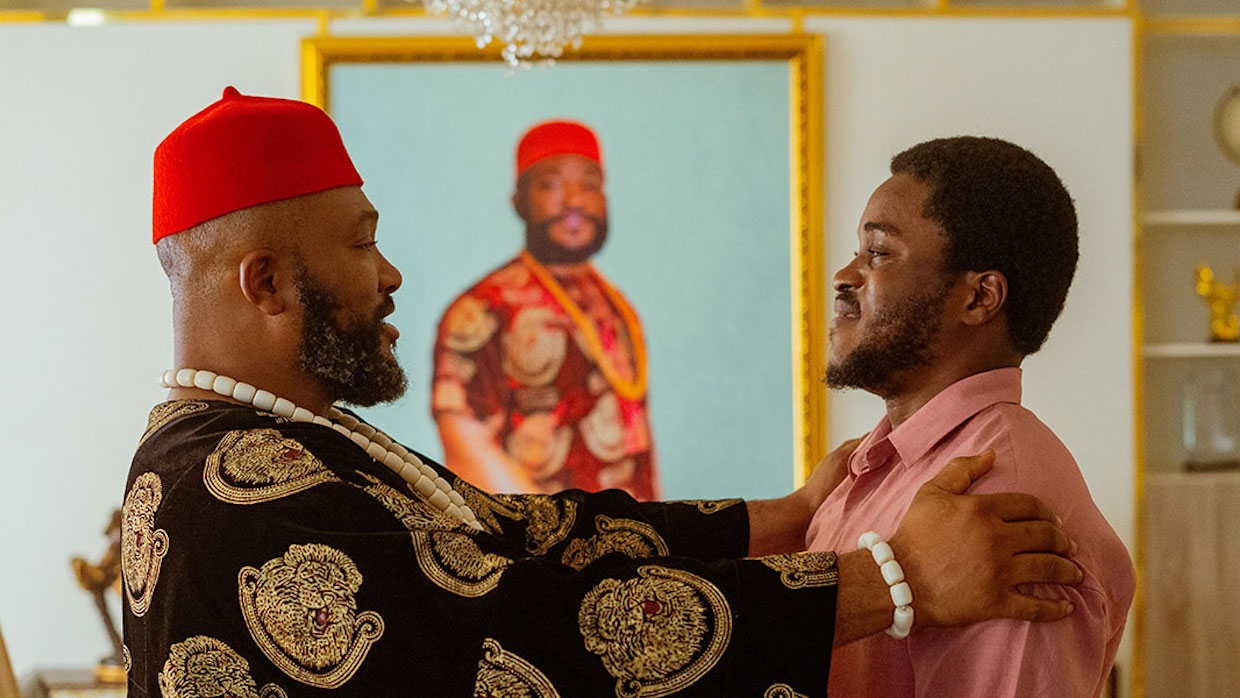 A Black man wearing traditional African garb holds a younger Black man's shoulders. They look into each other's eyes in front of a portrait of the older Black man when he was younger.
