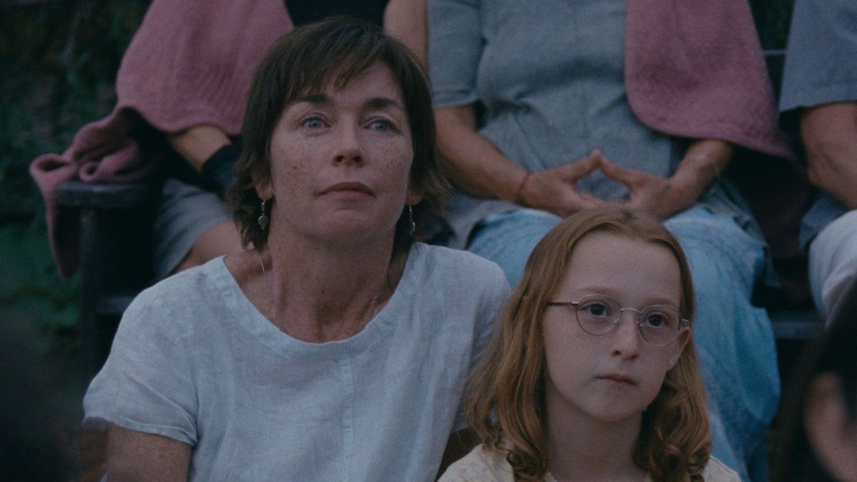 A woman in a white t-shirt sits behind a young girl wearing glasses.