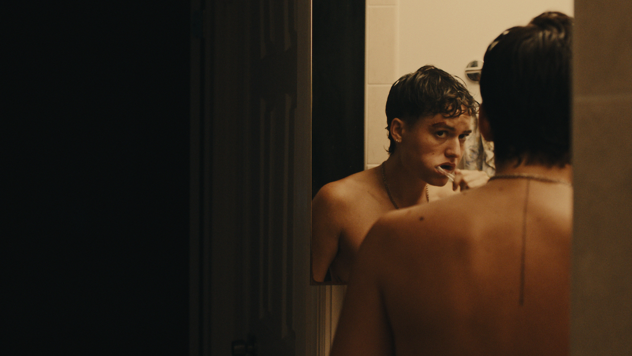 A trans man stands in front of a mirror shirtless brushing his teeth after a shower.
