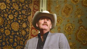 A man wearing a cowboy hat and grey suit jacket stands in front of two floral carpets hanging behind him.