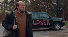 A balding, middle-aged man wearing a tan sweater and dark coat stands in front of a gray sedan with the word "loser" spray painted across the side in bright pink.
