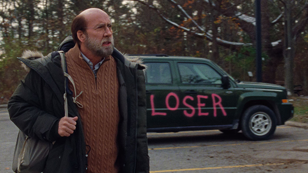 A balding, middle-aged man wearing a tan sweater and dark coat stands in front of a gray sedan with the word "loser" spray painted across the side in bright pink.