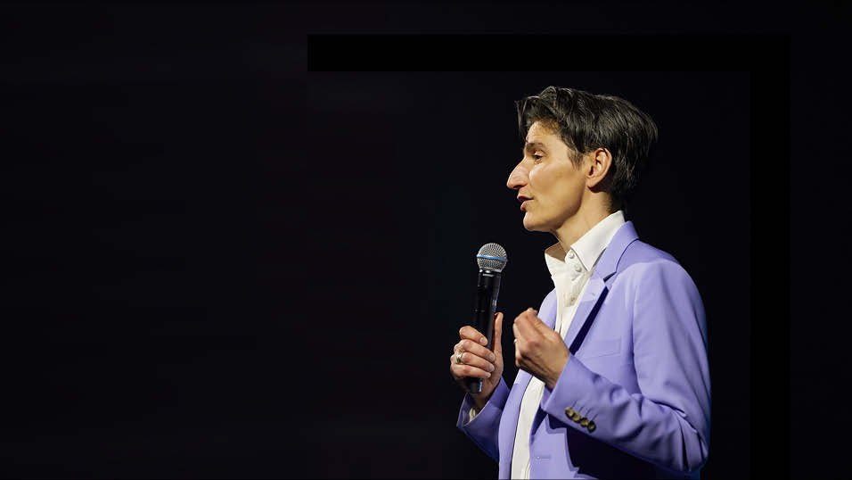 A 54-year-old woman with short dark hair, a purple suit and white shirt holding a microphone on a dark stage.