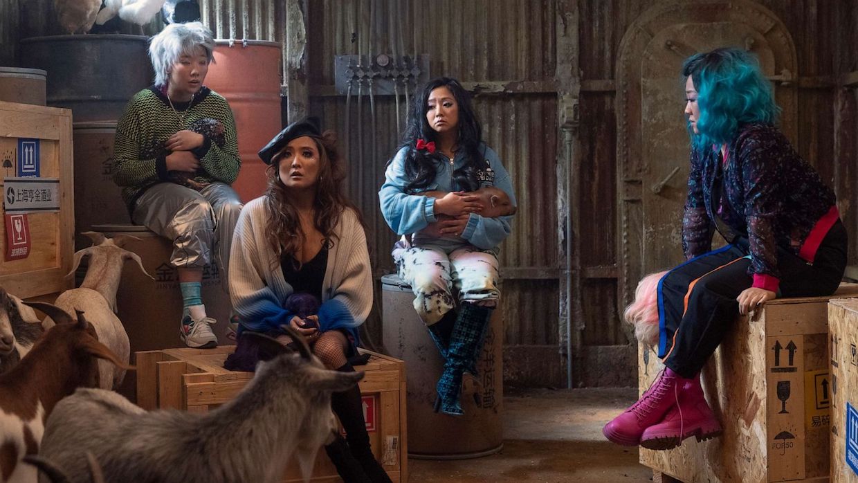 Four women sit in a storage room. They wear brightly colored clothes and accessories, but look mildly bewildered.