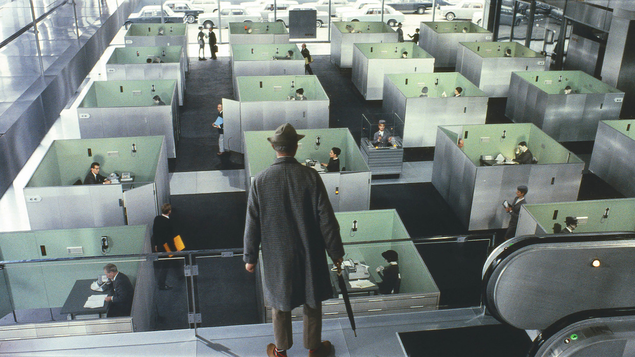A man wearing a hat and raincoat overlooks rows of symmetrical office cubicles below.
