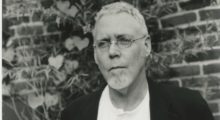 A grey-haired and bearded white man in his 70s wearing a black jacket and white t-shirt.