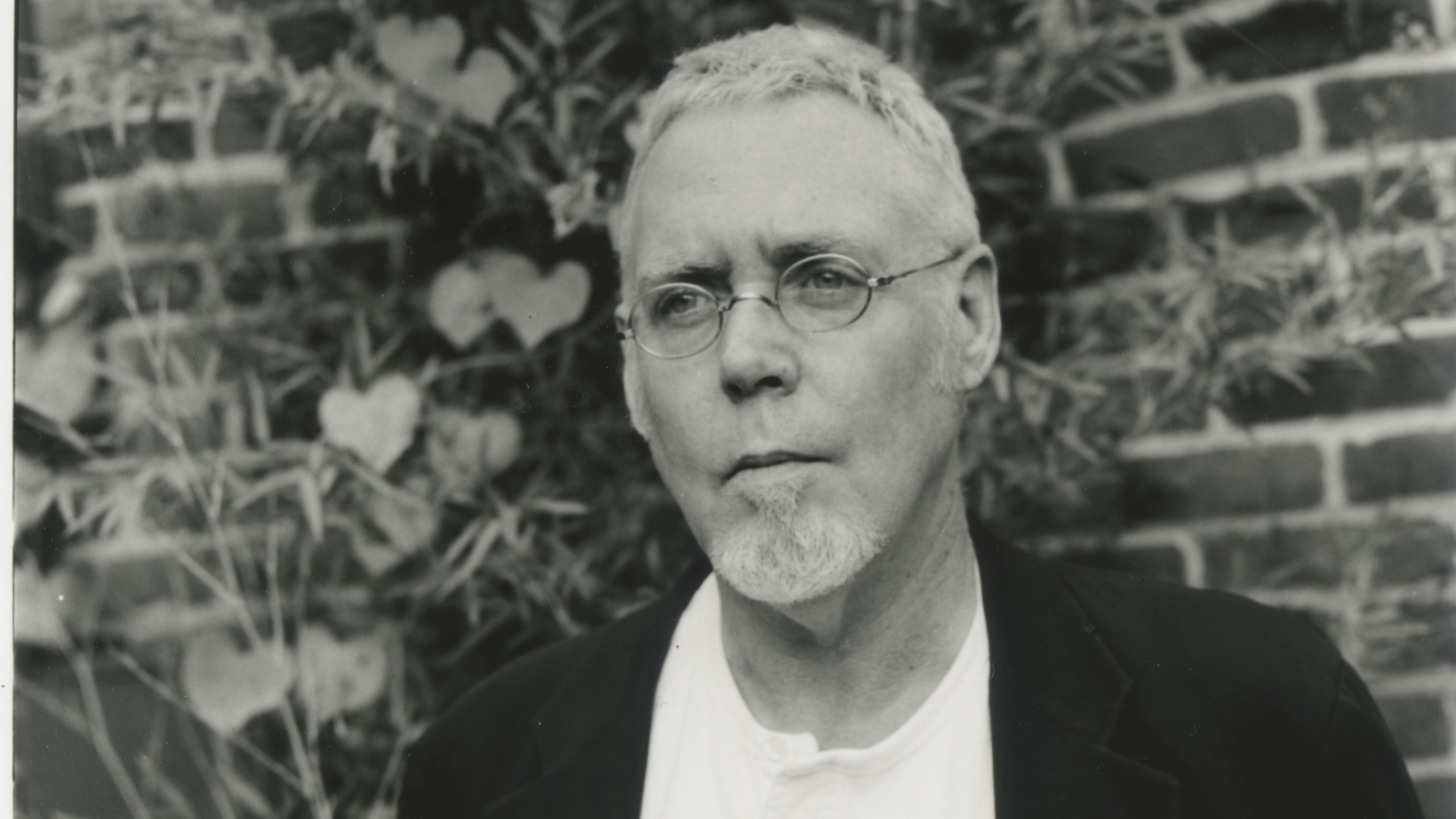 A grey-haired and bearded white man in his 70s wearing a black jacket and white t-shirt.