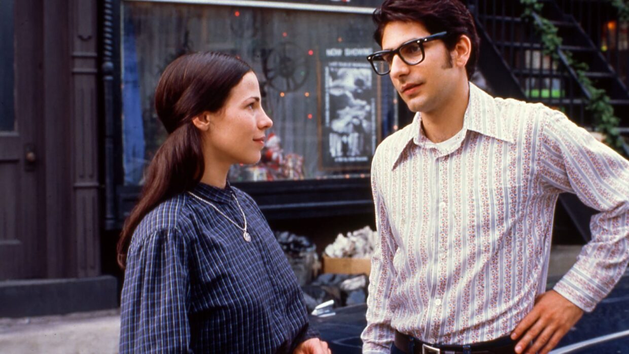 A young woman with long brown hair, blue dress and gold pendant and a taller young man with dark hair, a striped shirt and glasses.