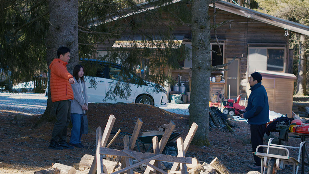 Takahashi (Ryuji Kosaka) points at a pile of wood alongside his colleague Mayuzumi (Ayaka Shibutani) and local resident Takumi (Hitoshi Omika) in Evil Does Not Exist