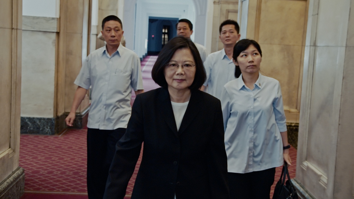A woman walks through a hallway in a dark blazer and white shirt while four people in light blue shirts follow behind her.