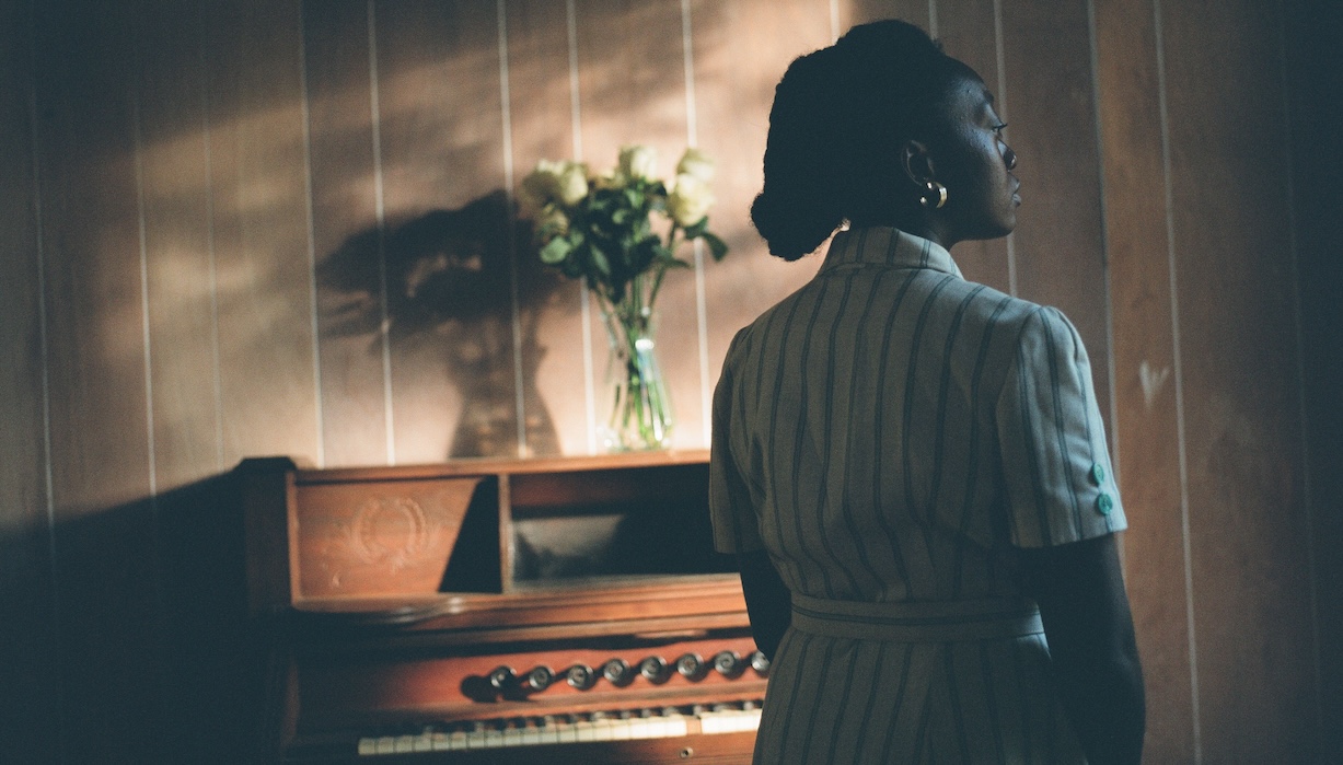 A woman stands in front of an upright piano with a vase of flowers resting on top of it.