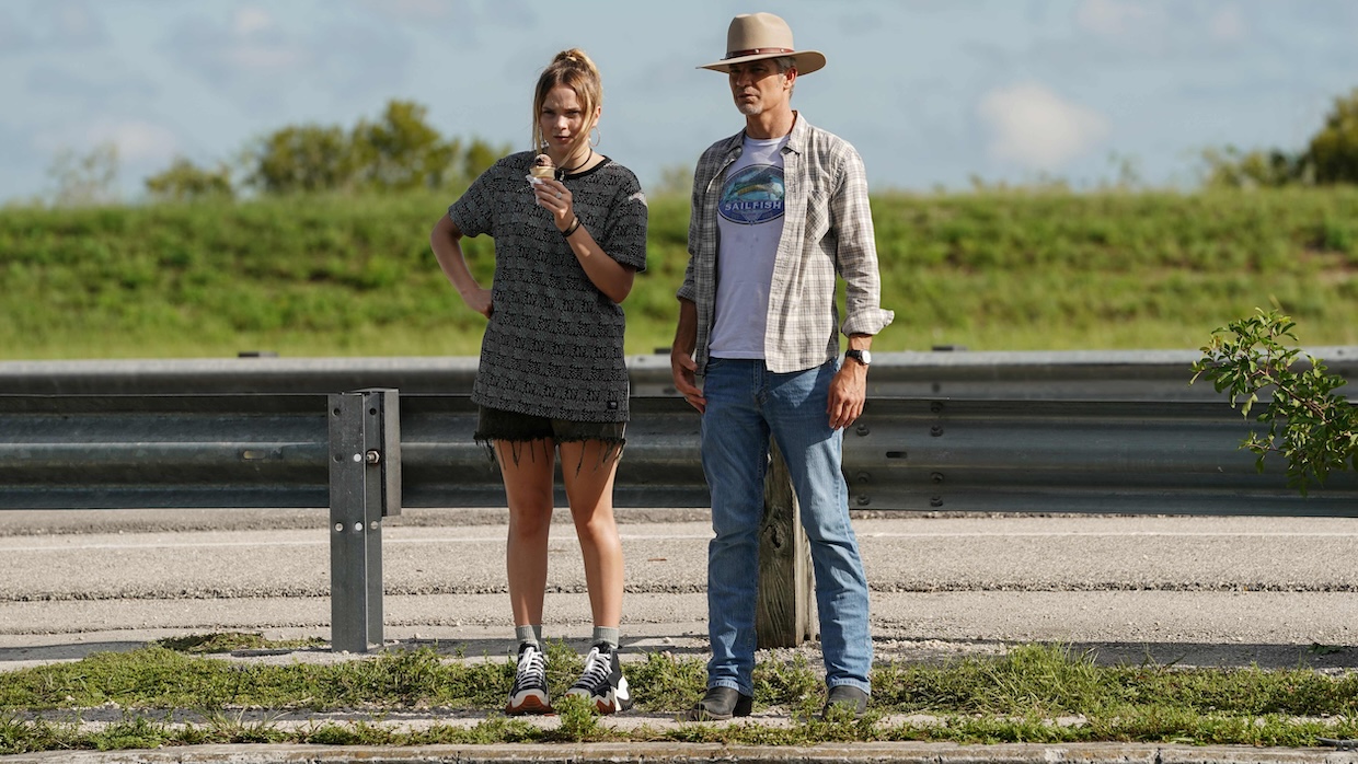 A teenage girl with an ice cream cone and a middle-aged man in a cowboy hat stand next to a freeway railing.