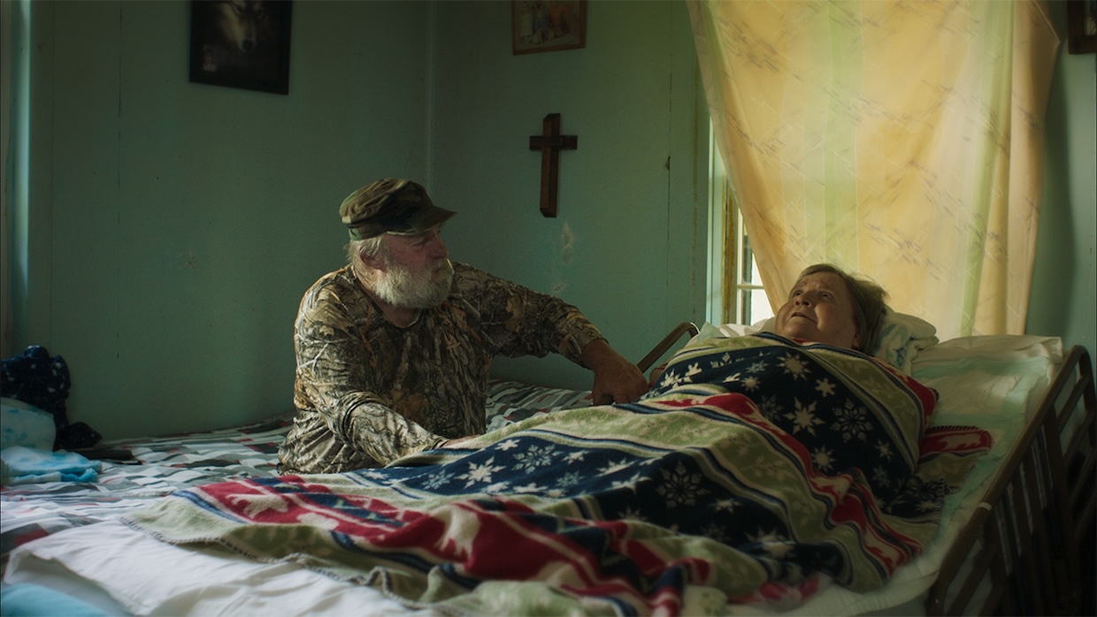 An older man sits at the bedside of a woman lying on a bed, with a crucifix on the wall between them.