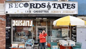 A man clutches his forehead standing beneath the awning of a record store.