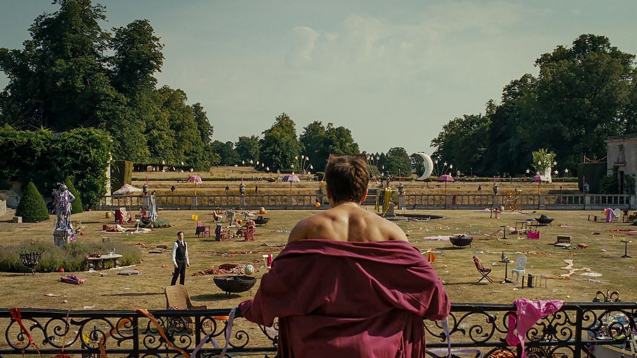 A man in a bathrobe stares out at a large trashed lawn littered with the remains of a party.