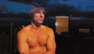 A shirtless man stares out a window at a cityscape