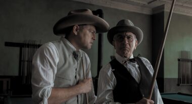 Two white men in cowboy hats stand next to each other. The one on the right holds a rifle.